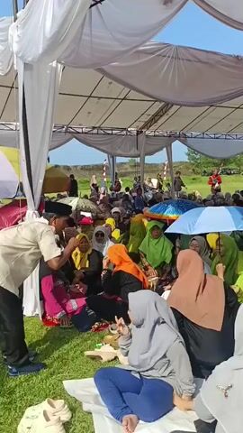 A low-angle shot captures a large group of people, predominantly women and children, gathered under a white tent-like structure on a grassy field. The tent’s frame is visible, revealing a blue sky above. Many of the women are wearing hijabs in various colors, including green, orange, grey, and brown. A few people are holding umbrellas for shade.  The group is sitting on the grass and on colorful carpets, some are eating. In the background, there are additional people standing and walking on the field, with trees visible on the horizon. The camera appears to be moving slightly.