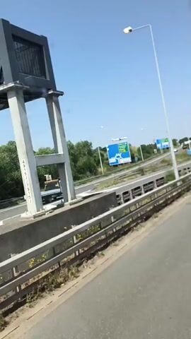 The video shows a view from a vehicle traveling on a highway in the Borough of Spelthorne, United Kingdom. The highway has multiple lanes, with traffic visible in the distance. There are large blue highway signs indicating destinations and distances, and a sign stating "No hard shoulder for 400 yds." The weather appears to be clear and sunny, with a bright blue sky. Trees and greenery line the sides of the highway, suggesting a rural or semi-rural setting. A truck and several cars are visible on the road. The time of day appears to be daytime, based on the bright sunlight and clear sky. There are no people clearly visible in the video, but the presence of vehicles indicates human activity. The overall mood is one of travel and transit.