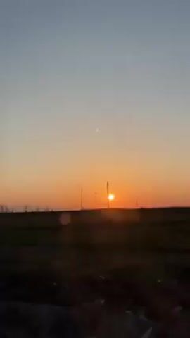 The video shows a sunset viewed from a moving vehicle. The sun is low on the horizon, appearing as a bright orange disk against a sky that transitions from light blue at the top to orange near the horizon. In the distance, there's a flat field with silhouettes of trees and multiple power line towers. The ground is visible at the bottom of the frame and shows rough terrain.