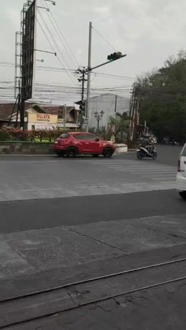 A red SUV drives through a crosswalk as a white SUV turns at an intersection. A motorcycle passes by, and a white car is partially visible on the right. Traffic lights are visible and green. A large billboard is on the left, and trees are on the right. The sky is overcast.