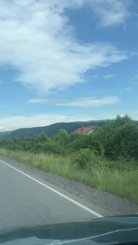 The video captures a daytime view from inside a moving vehicle traveling along a rural road in Nevytske, Ukraine. The asphalt road features a white solid line marking the shoulder. On the right side of the road, there is abundant green grass and dense trees, with more forested hills visible in the background. Several houses with red roofs are scattered among the trees in the distance. The sky is bright blue with numerous white, puffy clouds. Power poles line the side of the road. A white car is seen driving ahead in the distance. The scene suggests a pleasant drive through a green, hilly landscape under clear weather. No visible text is present.