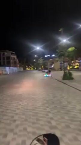At night, a child on a tricycle with glowing multicolored lights rides on a brick road next to a building under construction. A person in black walks next to the child. Overhead streetlights and lights from nearby businesses illuminate the otherwise dark area. There is signage and construction materials along the side of the road.