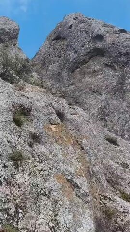The video shows a steep, rocky mountainside under a bright blue sky with scattered clouds. The camera angle is looking upwards, emphasizing the height and ruggedness of the terrain. Sparse green vegetation, including small bushes and grasses, clings to the rocks in various places. Towards the bottom right of the frame, a portion of a man-made structure, possibly a staircase or railing, is visible against the rock face. The rocks themselves are rough and textured, with variations in color ranging from light gray to reddish-brown. The overall impression is one of a natural, mountainous environment that has been partially accessed or modified by human construction.