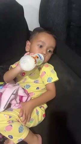 A young child wearing a yellow outfit with a fish pattern is sitting on a dark couch, drinking from a baby bottle. The child is holding the bottle with their right hand and a colorful blanket is draped over their lap. The bottle has a cartoon image of a tiger on it.