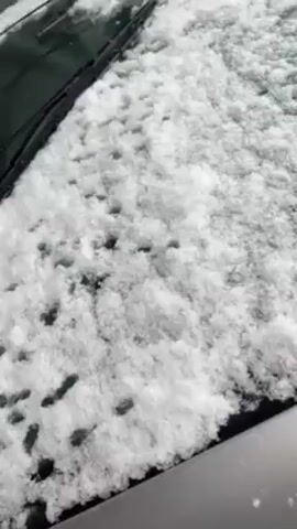 The video shows a car windshield covered in fresh snow. A hand reaches in and wipes some of the snow off the glass. The car appears to be silver in color. The snow is powdery and covers the entire windshield.