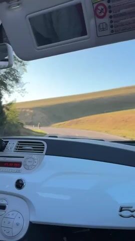 The video is filmed from inside a white car, looking out through the windshield. The car's dashboard, including a radio and a "500" logo, is visible in the foreground. The car appears to be moving along a winding road in a rural, hilly area. The sun is low in the sky, creating a bright glare and lens flare that obscures parts of the view. Green hills with dry grass and scattered trees are visible on either side of the road. In the distance, a person on a bicycle can be seen on the road. The sky is clear and blue. The timestamp indicates the video is approximately 4 seconds long.