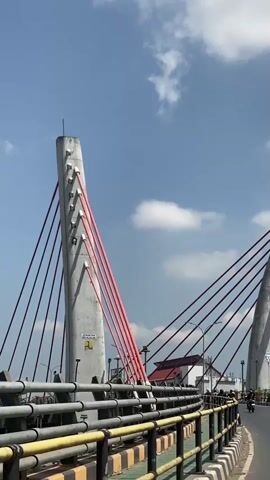 The video shows a view from a bridge with a clear blue sky and some clouds above. There are concrete pillars with red cables extending from them, characteristic of a cable-stayed bridge. A road runs across the bridge, with yellow and black striped barriers along the side. Several people are visible on motorcycles and a scooter, riding on the road. In the background, there are buildings and streetlights. The camera is moving forward, suggesting it is mounted on a vehicle or being panned by a person.