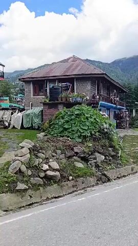 The video shows a vehicle driving along a paved road. On the left side of the road, there is a house with a red roof, stone walls, and a small garden. Further down the road, there are several other buildings and vehicles, including SUVs. The road is winding and curves to the right, with mountains covered in trees in the background. The sky is partly cloudy with blue patches. There are also signs visible along the road, one of which appears to be a medical center. The overall scene depicts a scenic mountain village or town.