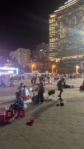 A wide shot shows a bustling outdoor plaza at night. A tall, illuminated building with a unique curved facade stands prominently in the background, its windows glowing with warm light. To the right of this building is a white, tulip-shaped structure, also illuminated. The plaza is paved with patterned tiles. Numerous people are scattered throughout the scene, some walking, others sitting on small plastic stools. Several individuals appear to be engaged in activities that suggest street vending or informal gathering, with bags, packages, and what could be food items visible on the ground. Motorcycles are parked in various locations, and a few appear to be in motion. The overall atmosphere is lively and active, characteristic of a public space at night.