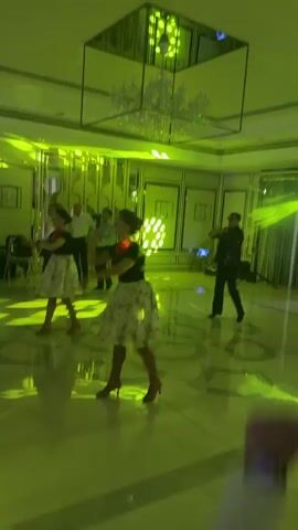 A group of people are dancing in a room with green lights. Two women in floral skirts and black tops are in the foreground, dancing in unison. A man in a dark suit is in the background, also dancing. Several other people are standing and watching. The room has a shiny floor and a large chandelier.