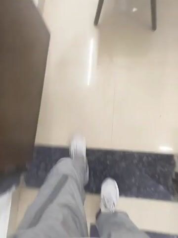 The video shows a first-person perspective of a person walking on a tiled floor in what appears to be an office or workspace. The person is wearing light gray pants and white sneakers. As they walk, the camera captures glimpses of office furniture including a wooden desk with various items on top such as cups and what appears to be cleaning supplies, a green wastebasket, and an office chair. The flooring is light-colored and shiny. The camera is shaky, reflecting the movement of the person walking.