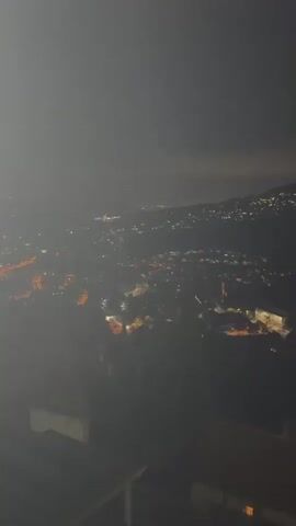 The video shows an aerial view of a city at night. The city lights are visible, creating a twinkling effect against the dark landscape. The camera appears to be moving slightly, giving a sense of depth and perspective as it pans across the scene. The lighting is dim, and the atmosphere is hazy, suggesting a slightly foggy or misty night. In the foreground, the edge of a structure, possibly a balcony or rooftop, is visible, adding a sense of being an observer. The overall impression is one of a quiet, sleeping city bathed in the glow of artificial lights.