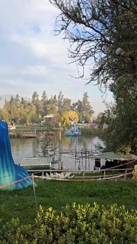 The camera pans over a tranquil lake featuring a colorful, bird-shaped sculpture at its center. The sculpture, which appears to be made of fabric and lights, is positioned on a floating platform, surrounded by additional floral and crystalline structures. Several people in kayaks paddle across the lake. In the background, trees line the shore, and a few small structures can be seen amidst the greenery. The foreground includes a grassy area, a wire fence, and a portion of a blue, inflatable structure.