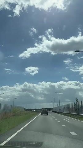 A car drives on a highway with a bright sunny sky and clouds above. The highway has multiple lanes and is lined with sound barriers. There are streetlights visible along the road.