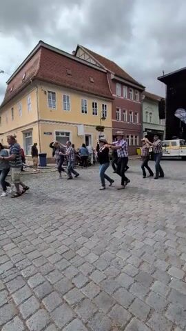 This video captures a group of people dancing in a cobblestone square, with buildings in the background. Several individuals, predominantly male, are wearing plaid shirts and cowboy hats, suggesting a country or western theme. They appear to be engaged in line dancing or a similar synchronized group dance. Some individuals are holding hands, moving in unison to unseen music. The environment suggests a town square with yellow and red buildings, possibly a European setting given the architecture. The sky is overcast. There are other people in the background, some seated and observing, while others stand near the edges of the scene. A white vehicle with yellow accents is visible on the right side, near a stage-like structure. The overall atmosphere seems festive and public.