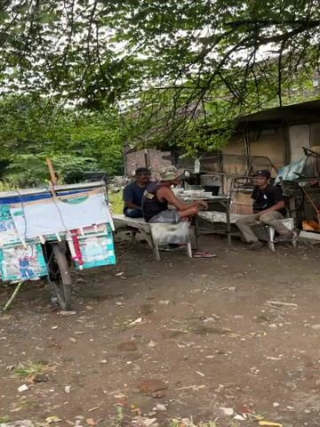 The video begins with a wide shot of a yard with two men sitting on chairs and talking under a tree. In the background, there are piles of scrap metal. The camera then pans to the right, revealing a motorcycle parked in front of a small building with a covered outdoor area. Inside the building, there are shelves stocked with goods. Further to the right, a red air compressor is visible outside the building. Another person is seen sitting on a bench to the left of the air compressor. The video ends with a shot of the front of the building, showing more shelves and items for sale.