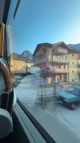 The video captures a view from a moving bus, looking out the window. The scene is an urban environment with buildings and mountains in the background. The sun is shining, and the sky is blue.

On the left side of the frame, the interior of the bus is visible. This includes a yellow curtain, a portion of a seat with a patterned fabric and a wooden armrest. The window itself shows reflections of the bus interior.

Outside the window, a street scene unfolds. There are several multi-story buildings with light-colored facades, balconies, and shutters. One building appears to be a gas station with an Esso sign. Vehicles, including a white car and what looks like a bus, are visible on the street. The mountains in the distance are rocky and steep, and the overall impression is that of a town situated in a mountainous region. The motion of the bus causes the outside scene to be slightly blurred, giving a sense of movement.