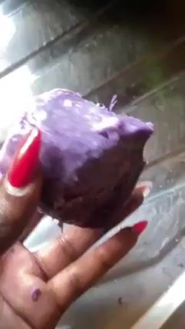 A person with dark skin and red nail polish is holding a piece of purple yam in their hand. The yam is cut in half and has a white center. The background is a stainless steel sink. The person rotates the yam to show the inside.