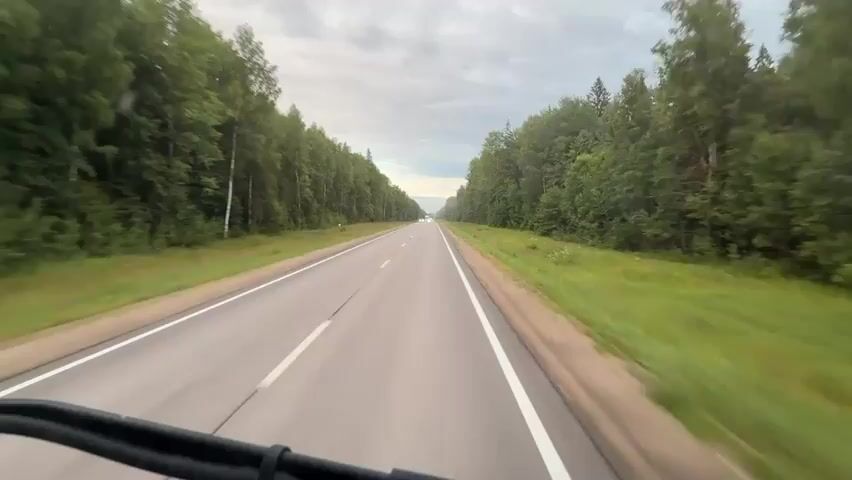 The video shows a point-of-view shot from a vehicle traveling on a two-lane road through a forest. The road is asphalt with white lane markings. Tall green trees line both sides of the road. The sky is cloudy. A white car is visible in the distance traveling in the opposite direction. Further down the road, a white truck with a trailer is traveling in the same direction as the camera. The camera is positioned low, showing a portion of the vehicle's interior, possibly a dashboard or windshield frame, at the bottom of the frame. The video appears to be a short clip of a road trip.