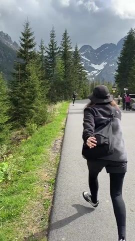 The video shows a young woman with medium brown hair, wearing a black hat, a black jacket over a light gray top, black leggings, and black sneakers, doing a skipping dance on an asphalt road. She is facing toward a mountain range partially covered with snow under a cloudy sky. There are tall green evergreen trees to her left, and more people and a white vehicle are visible further down the road to her right. She is also carrying a bag over her shoulder.
