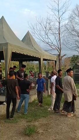 This is a video taken outdoors during the day, likely in the late afternoon or early evening, given the lighting. The scene appears to be a public gathering or event.

In the first frame, a group of men are standing around a temporary tent structure. They are dressed in casual clothing, including t-shirts and jeans. Some men are looking towards the tent, while others are talking to each other. The ground is dirt and grass.

The subsequent frames show a large stage set up for an event. The stage is decorated with green banners and a large LED screen displaying the words "GEMA MUHARRAM". There are lights and sound equipment on the stage, and several people are visible on and around it, possibly setting up or preparing for a performance. Several motorcycles are parked in front of the stage. The overall atmosphere suggests an outdoor festival or celebration.