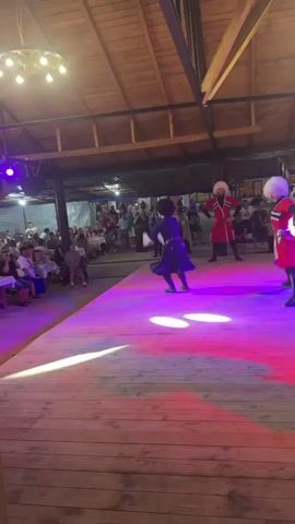 The video captures a traditional dance performance taking place on a wooden stage illuminated by purple and pink lights. Three dancers are prominently featured, all wearing elaborate traditional costumes. One dancer, in the center and slightly elevated, is mid-jump, dressed in a dark outfit with intricate details and a distinctive hat. To the right, a dancer in a vibrant red costume with a white fluffy hat is in a dynamic pose, her skirt swirling. Another dancer in a similar red costume and white hat stands behind her, looking towards the audience. Several other individuals in similar attire are also visible in the background, some appearing to be part of the performance or an accompanying ensemble.

The setting appears to be an outdoor or semi-outdoor venue, with a wooden roof structure overhead and visible lighting fixtures. In the background, an audience is seated at tables, observing the performance. The overall atmosphere suggests a lively cultural event or celebration. The lighting casts a theatrical glow on the stage, enhancing the visual spectacle of the dance. The time of day is likely evening or night, given the artificial lighting and the presence of spotlights. The location is identified as Durypsh, Abkhazia, hinting at a performance of traditional Abkhazian dance. The dancers' poses and movements convey energy and skill, suggesting a high-spirited and engaging performance. No visible text is present in the video.