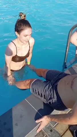The media file shows a woman in a brown bikini in a swimming pool, holding onto the legs of a person lying on the edge of the pool. The woman is smiling and appears to be in a playful mood. The person on the edge of the pool is wearing grey shorts with the word "JORDAN" visible on the leg. The water in the pool is blue and appears to be clear. There is a metal ladder to the right of the frame, suggesting a public pool or a well-maintained private pool. The lighting suggests it is daytime, likely sunny. The scene is set at a pool, possibly in Vladimirovka, Moldova. The overall mood seems lighthearted and fun.