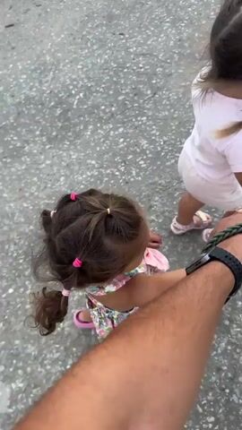 The video shows a point-of-view perspective from a person walking with two young children and a dog on a leash. The first child, in the foreground, is wearing a pink and floral dress and has her hair in multiple ponytails with pink ties. She is holding the person's hand. The second child, slightly further ahead, is wearing a white t-shirt and white shorts, and her hair is in two braids with purple ties. She is also holding onto a leash connected to a dog. The dog is a medium-sized dog with a merle coat pattern, wearing a chain collar. The third person, presumably the one filming, is holding the leash with their right hand, which is wearing a black smartwatch. The ground appears to be a paved or asphalt surface with some gravel. There are trees and vegetation on the sides of the path, suggesting an outdoor environment, possibly a rural or suburban road. The lighting indicates it might be early morning or late afternoon. The video captures the act of walking and the interaction between the children, the dog, and the person.