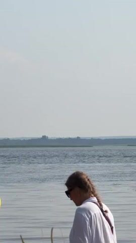 A woman with braided hair and sunglasses, wearing a white shirt, stands near a body of water. In the background, a white motorboat with two people wearing orange life jackets speeds across the water. A man in a bright yellow shirt is on the left, talking on his phone. Tall reeds are in the foreground. The sky is hazy and bright.