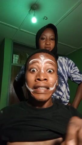 Two people are visible in the video, framed in a selfie. The person in the foreground is a dark-skinned man with short hair, wearing a black shirt. His face is partially covered in white makeup, forming a tribal-like pattern. He is making exaggerated facial expressions, including widening his eyes and baring his teeth.  Behind him is a woman wearing a black hijab and a blue and white tie-dye shirt. The woman smiles and gives a thumbs up. They appear to be indoors. The room has a green wall, a white ceiling with a single, lit light bulb. The video appears to be taken with the front facing camera. There is loud music playing in the background.