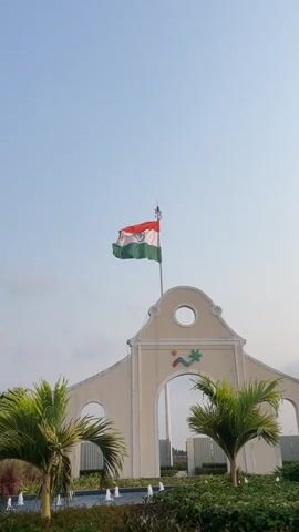 The video shows the Indian flag waving atop a white archway structure, with a clear blue sky in the background. The flag is brightly colored with the traditional saffron, white, and green stripes, and the Ashoka Chakra is visible in the center. The archway has a decorative design, including a circular opening at the top and a company logo near the apex of the arch. Flanking the archway are two small palm trees and a water feature with multiple fountains. The camera remains static, providing a clear, steady view of the scene.