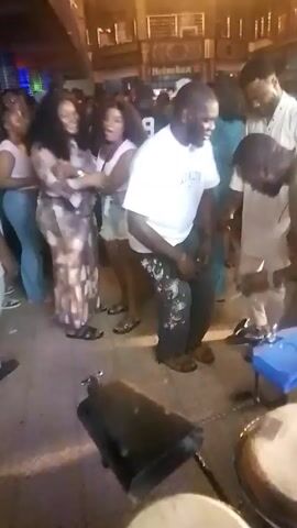 The video shows a group of people dancing and socializing at what appears to be a club or outdoor venue. The scene is dimly lit by string lights, creating a warm ambiance. There are several individuals dressed in casual attire, including a man in a white shirt and patterned pants who is seen actively dancing. Others are engaged in conversations or moving to the music. The setting seems to be an open space with a bar or elevated platform in the background. The video is shot handheld, resulting in some movement and slightly shaky footage. The atmosphere seems lively and festive.