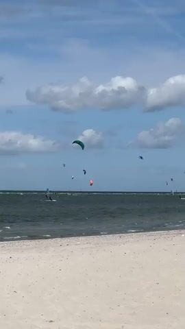 A low-angle, static shot looks out over a sandy beach towards a body of water under a bright, cloudy blue sky. Several kitesurfers are visible on the water, their colorful kites dotting the horizon. The waves are moderately sized, and the water appears choppy. The sand in the foreground is light brown and appears somewhat coarse. No individuals are clearly visible on the beach, only a vague, dark shape near the left edge that might be a person. The kitesurfers are dispersed across the water, some appearing to be closer to the shore than others. The scene conveys a sense of outdoor recreation and activity on a breezy day.