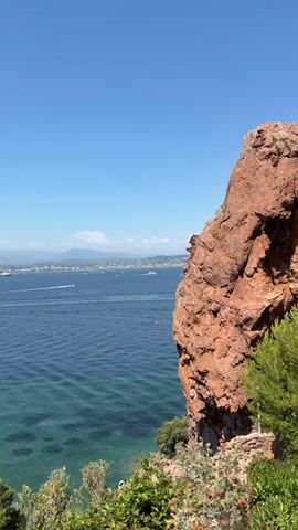 A vertical video shows a coastal scene from a high vantage point. In the foreground, a large, reddish-brown rock formation juts out from the right side of the frame, covered with some sparse vegetation. Below the rock, lush green bushes and trees are visible, with a small, pebbly beach at the water's edge. To the left, the clear, blue water of the sea stretches out, with patches of lighter turquoise indicating shallows. Several boats, including a large white yacht and smaller speedboats, are scattered across the water. In the distance, a coastline with hills and buildings can be seen under a bright blue sky with a few wispy clouds. The overall impression is a sunny day with calm seas and a scenic coastal view. There are a few people visible on the beach and in the water.