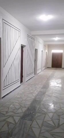 The video shows a long, brightly lit hallway. The walls are white, and there are dark brown doors along one side, set within decorative frames with black lines and a diagonal stripe pattern. The floor is tiled with a geometric pattern of light and dark gray. A large, dark wooden door is at the far end of the hallway. The camera moves slowly forward through the hallway.