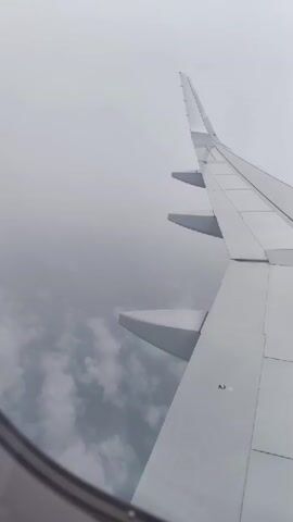 The video shows an aerial view from the window of an airplane. The wing of the plane is prominently visible, extending upwards and to the right of the frame. Below the wing, clouds are visible, appearing as a gray, textured layer. The sky is overcast and appears a uniform gray. The video is a still frame.