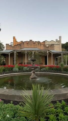 A vertical, wide shot shows a tranquil garden scene with a fountain in the foreground. The fountain has two tiers and is actively spraying water into a circular pond. Lush green plants and red flowers surround the pond. Behind the pond, a light-colored building with a verandah and crenellated parapets stands under a clear sky. The building appears to be an older structure, possibly a historic house or a garden pavilion. The overall atmosphere is peaceful and serene, with the gentle sound of the fountain.
