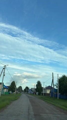 This media file depicts a rural street scene in Аргамак, Russia, under a bright blue sky filled with wispy white clouds. The perspective is from a vehicle moving down a paved road that shows signs of wear and tear.

On either side of the road, there are scattered houses with colorful roofs, typical of a village setting. Trees line the street, and utility poles with numerous wires crisscross overhead, creating a sense of established infrastructure. Some houses have fences and appear to be well-maintained, while others suggest a more rustic existence.

In the distance, a few people can be seen walking along the road, indicating the presence of life and activity within the community. The overall atmosphere appears peaceful and serene, suggesting a quiet summer afternoon. The sunlight, casting soft shadows, indicates it is likely late afternoon or early evening. The clear sky with scattered clouds points to fair weather. The scene evokes a sense of calm and the simplicity of rural life. No visible text is present in the media file.