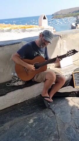 A man with a beard, wearing a cap, a dark t-shirt, and shorts, sits on a stone ledge, playing an acoustic guitar. He is facing to his right, looking down at the guitar. To his right, a black guitar case rests on the ground, next to a small blackboard with writing on it. Behind him, the blue sea stretches out, dotted with boats. A white building with a conical roof is visible on a hill overlooking the water. The man is wearing flip-flops. He appears to be a street performer.