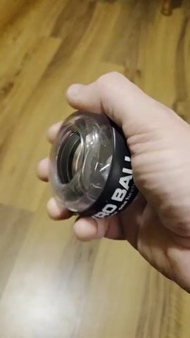 A hand holds a clear and black gyro ball over a wooden floor. The hand rotates the ball, which causes the internal gyroscope to spin rapidly. The words "GYRO BALL" are printed on the side of the ball.