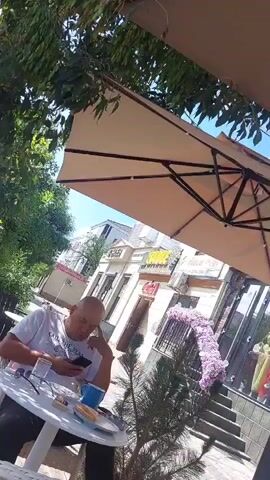 The video begins with a man sitting at a round table outside a cafe, looking at his phone. He is wearing a white t-shirt and black pants. On the table, there is a coffee cup, a plate with food, a cigarette pack, and a cell phone. A large umbrella provides shade over the table. The background shows a street with buildings, trees, and a purple floral decoration. The camera then pans to the right, showing the facade of the buildings across the street, with large windows and architectural details. Some reflections are visible in the windows. The paving stones of the outdoor seating area are also visible. The scene appears to be a casual outdoor cafe setting on a sunny day.