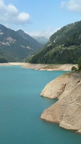The video shows a vast, turquoise lake nestled between towering, tree-covered mountains. The sky is partly cloudy. In the foreground, a steep, rocky embankment slopes down to the water's edge. A few small, orange buoys are visible in the lake, marking a boundary or hazard. The water level appears to be low, exposing a dry, tiered section of the embankment. A bird is captured mid-flight over the water. The scene is serene and natural, with the focus on the grandeur of the landscape and the striking color of the water.