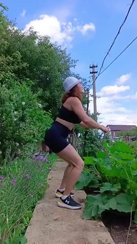 A woman in athletic wear and a white hat performs squats outdoors. She stands on a paved pathway surrounded by lush green foliage and purple flowers. The sky is blue with scattered clouds, and a utility pole with wires is visible in the background. The woman starts in a standing position, then lowers herself into a squat with her arms extended forward, and returns to a standing position. This action repeats throughout the short video clip.