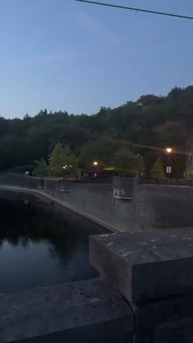 This video shows a night view of a stone church with illuminated windows and a bell tower. The church is located next to a cobblestone sidewalk and a street. In the background, there is a wooded hillside and a few streetlights. The sky is dark blue with a few faint contrails. The video appears to be filmed from a bridge or elevated vantage point overlooking the scene.