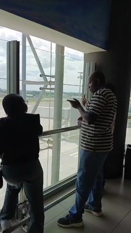 A woman and a man stand at a large glass window in what appears to be an airport, looking out onto the runway. The woman is wearing a dark jacket and jeans, with a small bag over her shoulder. The man is wearing a striped shirt and jeans, and he is pointing out towards the runway while holding something in his other hand. There is another person visible in the background, also looking out the window. The airport environment is evident through the runway and the overall architecture of the building.