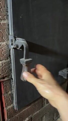 The video begins with a close-up of a person's hand holding a dark metal handle attached to a hinged metal door. The door is partially open, revealing a dark, interior space. The door is surrounded by brickwork. The hand turns the handle, and the door swings open further, revealing the interior of a fireplace or similar structure. The interior appears to be coated with soot and ash, with some debris visible at the bottom. A pink object, possibly a small bucket or container, is then inserted into the opening. The video pans slightly to the right, showing more of the brickwork and some wooden surfaces. The interior of the fireplace is fully visible at this point, and the pink object is no longer in view.