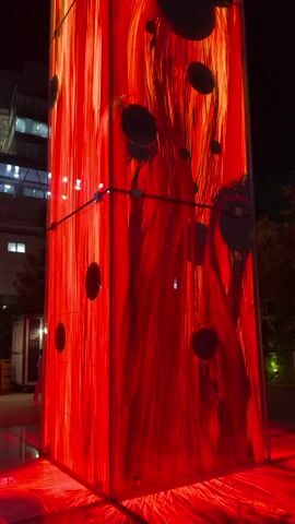 The video shows a tall, translucent tower illuminated from within by a vibrant, swirling red light. The light effect mimics the appearance of flowing lava or strands of fiery hair, with darker circular shapes interspersed throughout. The tower is rectangular and appears to be made of glass or a similar transparent material. It is situated outdoors at night, with glimpses of buildings and trees in the background, suggesting an urban or public space. The camera pans upwards, focusing on the dynamic light display within the tower. No people or animals are clearly visible in the frame.