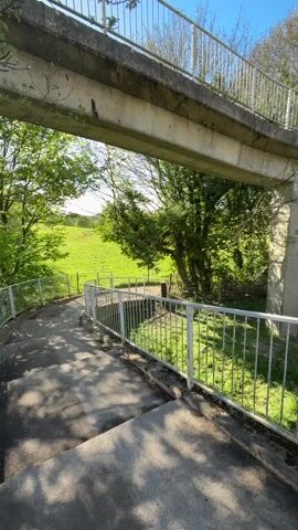 The camera, positioned at ground level, looks up at a concrete pedestrian overpass with white metal railings.  The overpass curves to the right and extends over a green field. Sunlight creates shadows on the stairs and surrounding surfaces. Two figures are walking across the overpass from left to right. The sky is blue with scattered clouds. The environment includes green trees and grass.