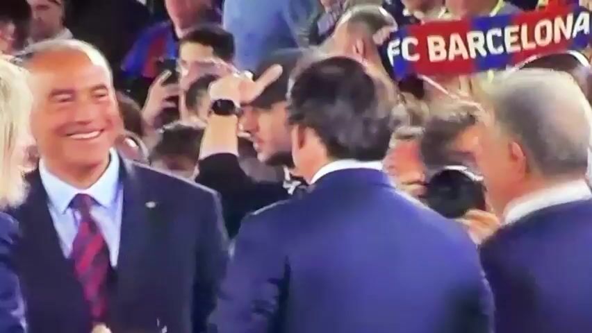 The video shows a group of people in a crowded setting. Several men in suits are visible, one of whom is smiling broadly. This man is wearing a dark suit, light blue shirt, and a red and black patterned tie. He hugs another man in a similar suit. The surrounding crowd includes people of diverse ages and ethnicities. Many are looking in the direction of the men in suits, and some are taking photos or videos with their phones. A "FC BARCELONA" sign can be seen in the background, suggesting the event may be related to the football club. The lighting is bright and suggests an indoor environment.