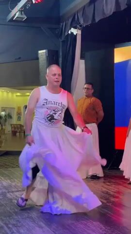 A bald man in a white tank top and a long, flowing white skirt is dancing on a stage. He is spinning and twirling, making the skirt flare out. To his right, another man in a patterned shirt and a similar white skirt is watching him. In the background, there is a large screen displaying the Russian flag, stage lighting, and some audience members. The overall environment appears to be a performance space or event venue.